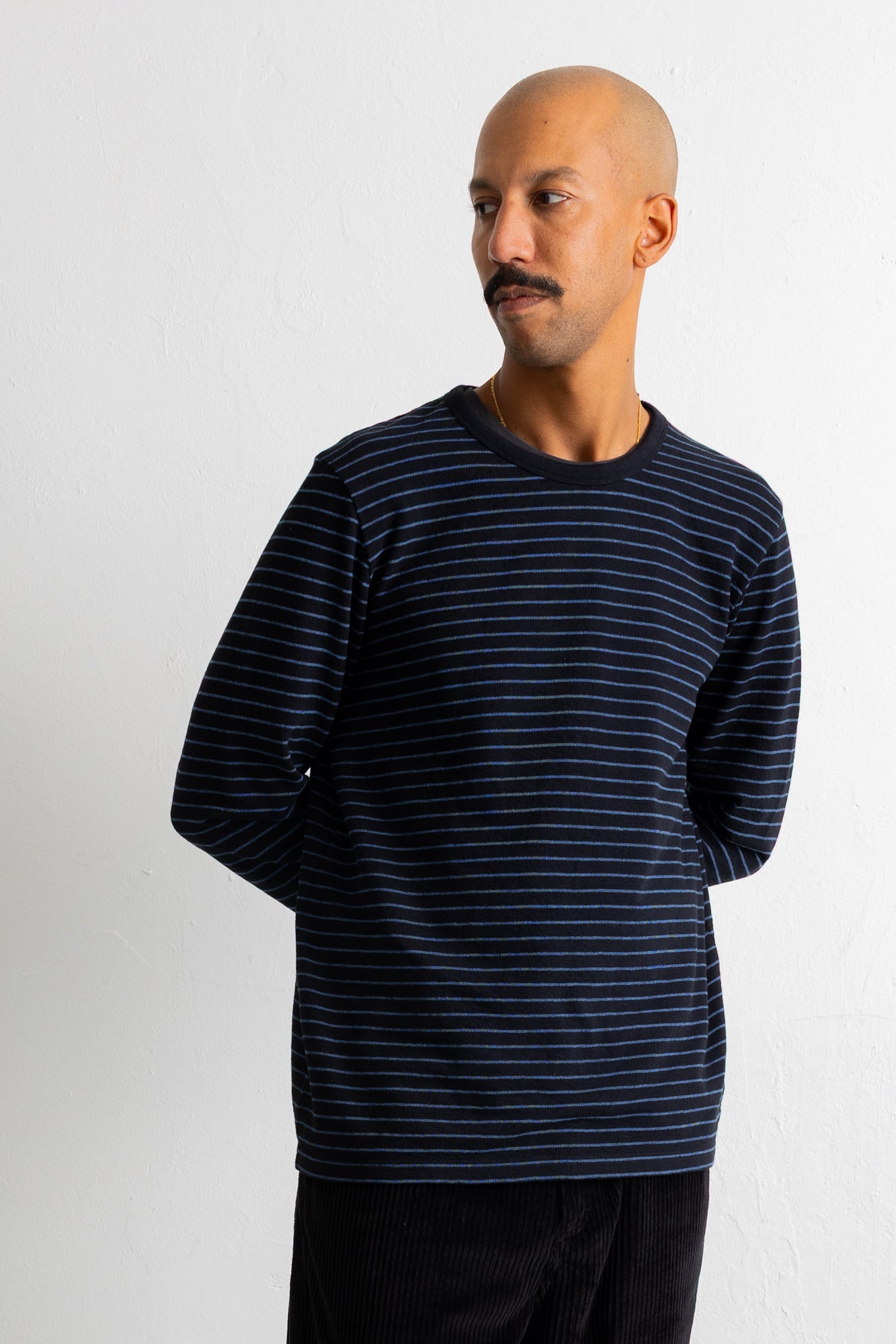 A man wearing a navy blue long-sleeve shirt with thin white horizontal stripes, standing against a plain light gray wall.
