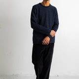 A man wearing a navy blue and black horizontally striped long-sleeve shirt, black pants, and black-and-white sneakers, standing against a white studio wall.