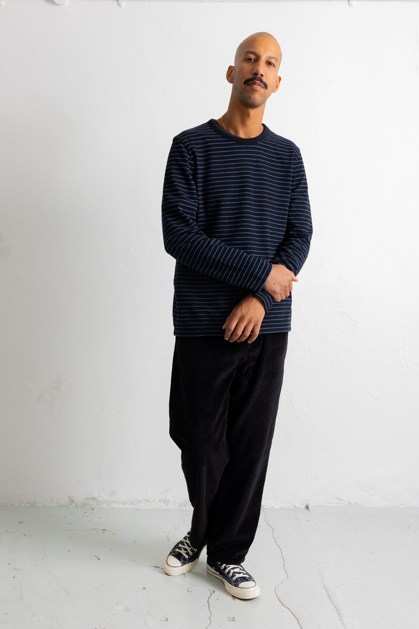 A man wearing a navy blue and black horizontally striped long-sleeve shirt, black pants, and black-and-white sneakers, standing against a white studio wall.