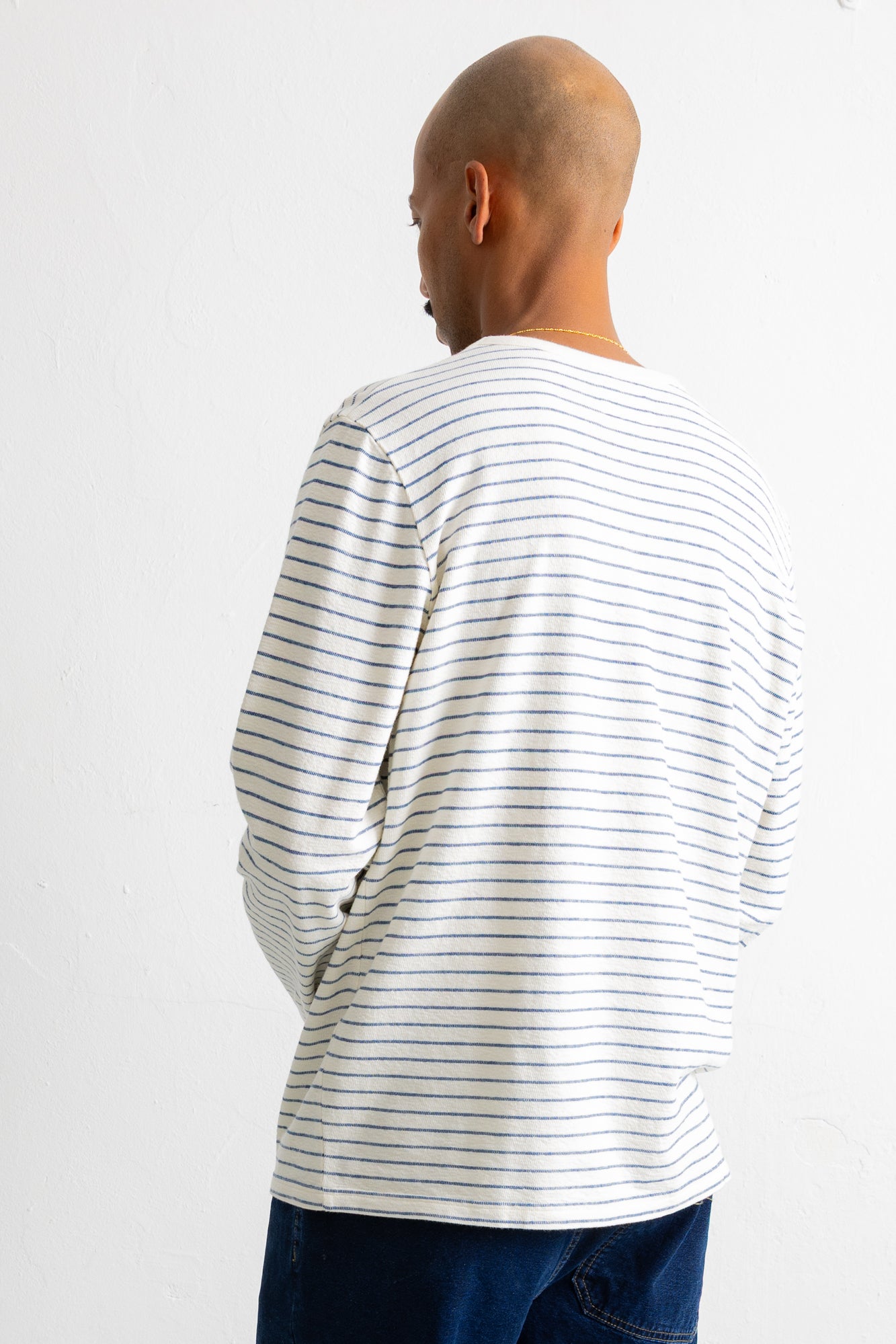 A man viewed from behind wearing a white long-sleeve t-shirt with thin dark horizontal stripes and dark blue jeans, set against a plain white background.