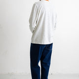 A man viewed from behind wearing a white long-sleeve t-shirt with thin dark horizontal stripes, dark blue jeans, and black canvas sneakers, standing against a white wall.