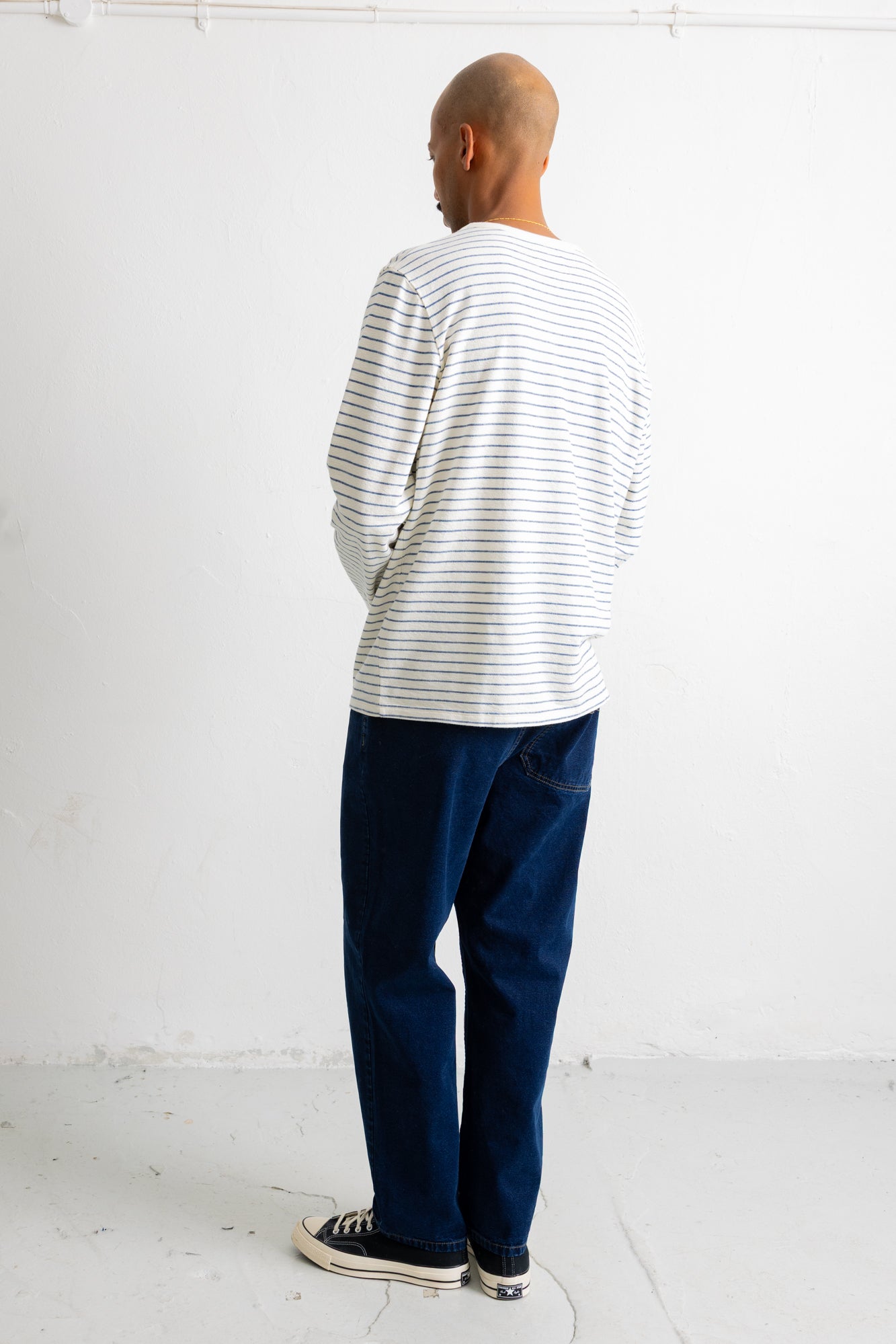 A man viewed from behind wearing a white long-sleeve t-shirt with thin dark horizontal stripes, dark blue jeans, and black canvas sneakers, standing against a white wall.