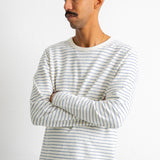 A man wearing a white long-sleeve crewneck shirt with thin dark horizontal stripes, paired with dark blue jeans, standing against a plain white background.