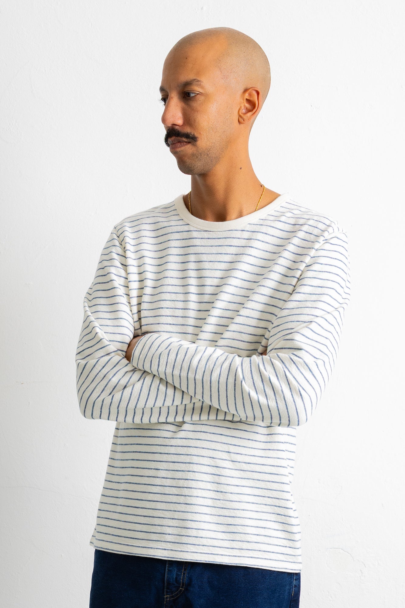 A man wearing a white long-sleeve crewneck shirt with thin dark horizontal stripes, paired with dark blue jeans, standing against a plain white background.