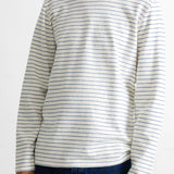 A man wearing a long-sleeve crewneck shirt with thin horizontal navy blue stripes on an off-white base, paired with dark blue jeans.