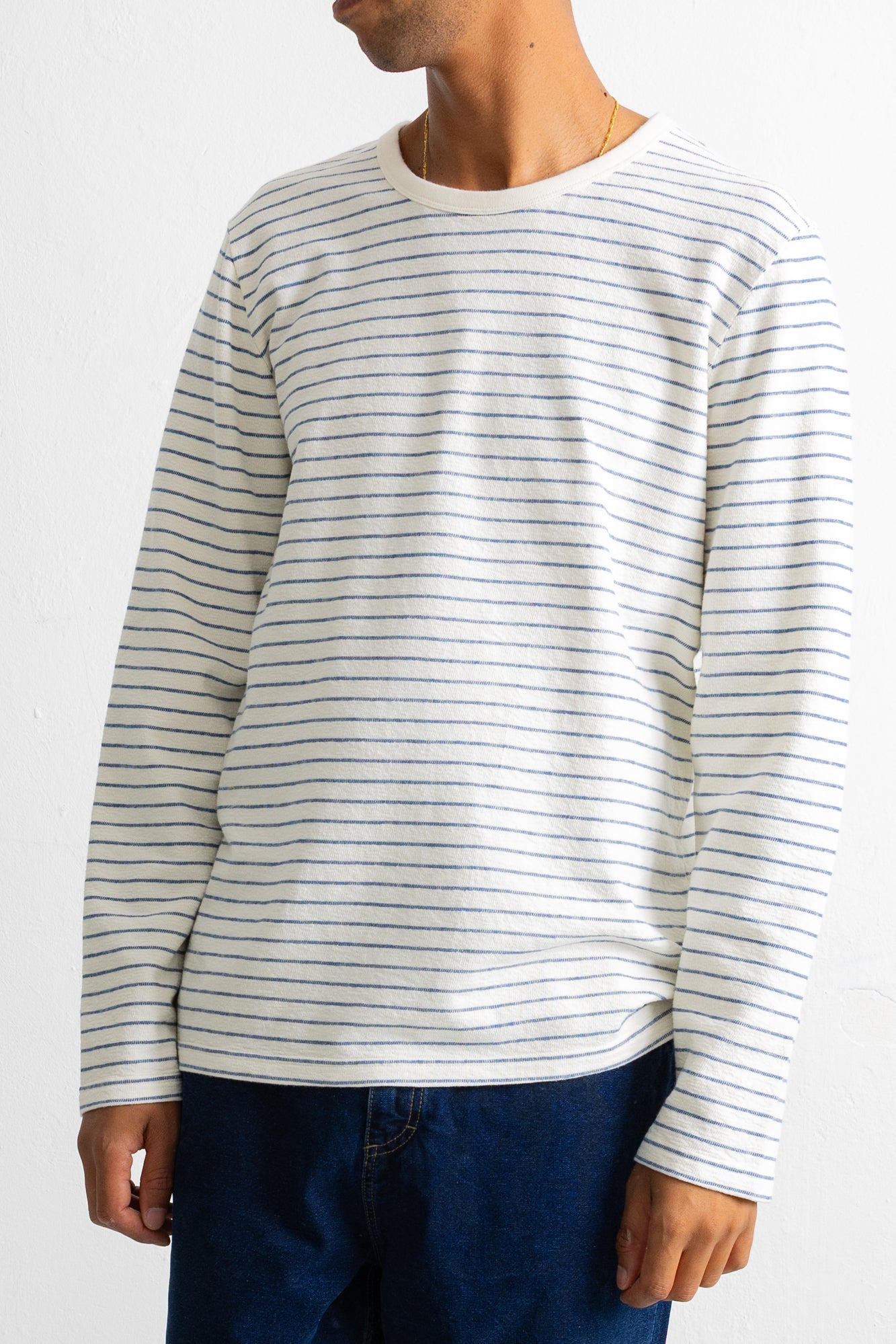 A man wearing a long-sleeve crewneck shirt with thin horizontal navy blue stripes on an off-white base, paired with dark blue jeans.