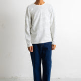 A man wearing a white long-sleeve crewneck shirt with thin gray horizontal stripes, paired with dark blue jeans and black-and-white sneakers, standing against a plain white wall.