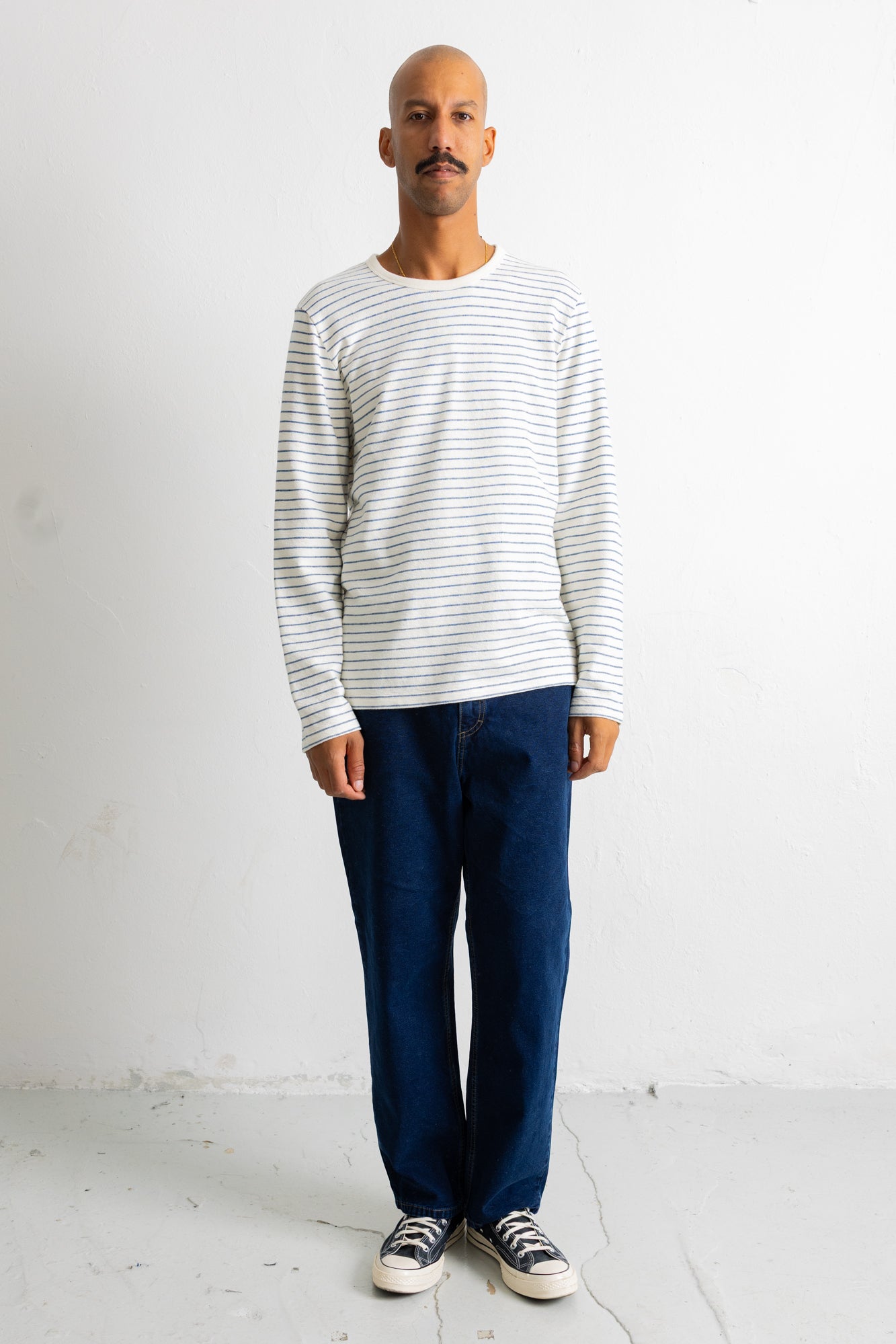 A man wearing a white long-sleeve crewneck shirt with thin gray horizontal stripes, paired with dark blue jeans and black-and-white sneakers, standing against a plain white wall.