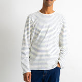 A man wearing a white long-sleeve crewneck shirt with thin dark horizontal stripes and dark blue jeans, standing against a plain light gray background.