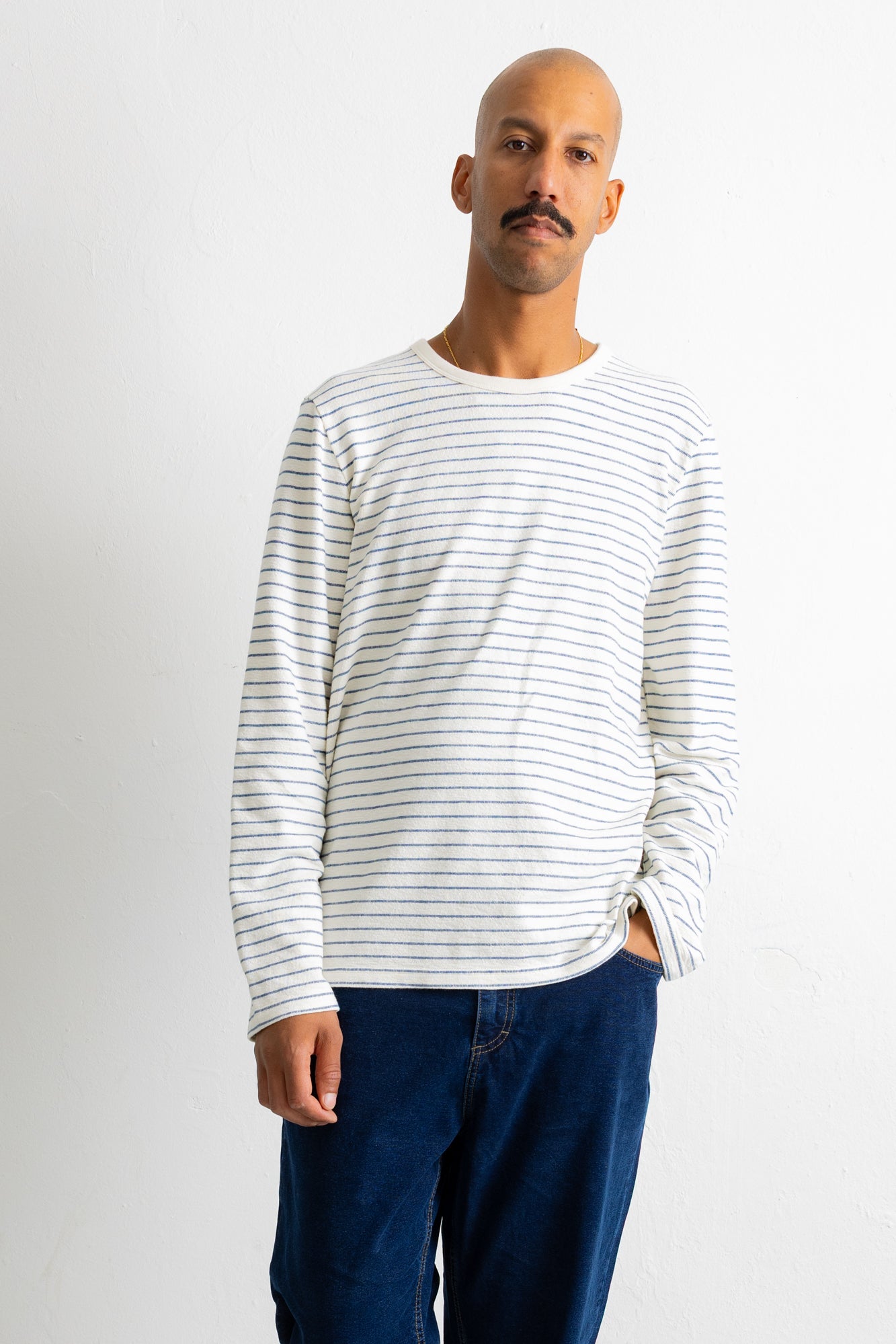 A man wearing a white long-sleeve crewneck shirt with thin dark horizontal stripes and dark blue jeans, standing against a plain light gray background.