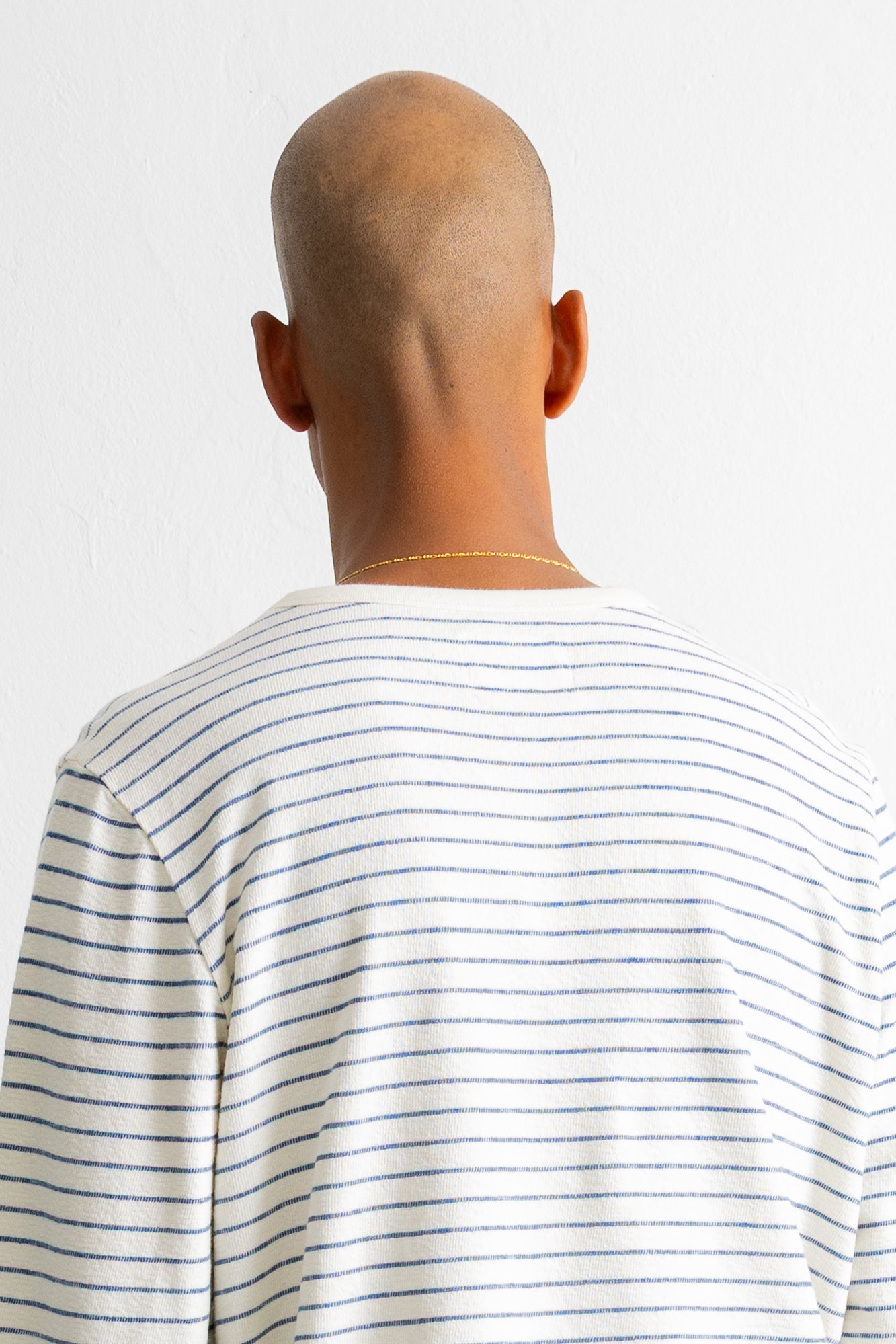 Rear view of a bald man wearing a white long-sleeve t-shirt with thin blue horizontal stripes, set against a plain white background.