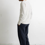 A man wearing a white long-sleeve shirt with thin black horizontal stripes and dark navy wide-leg trousers, standing in profile against a white wall.