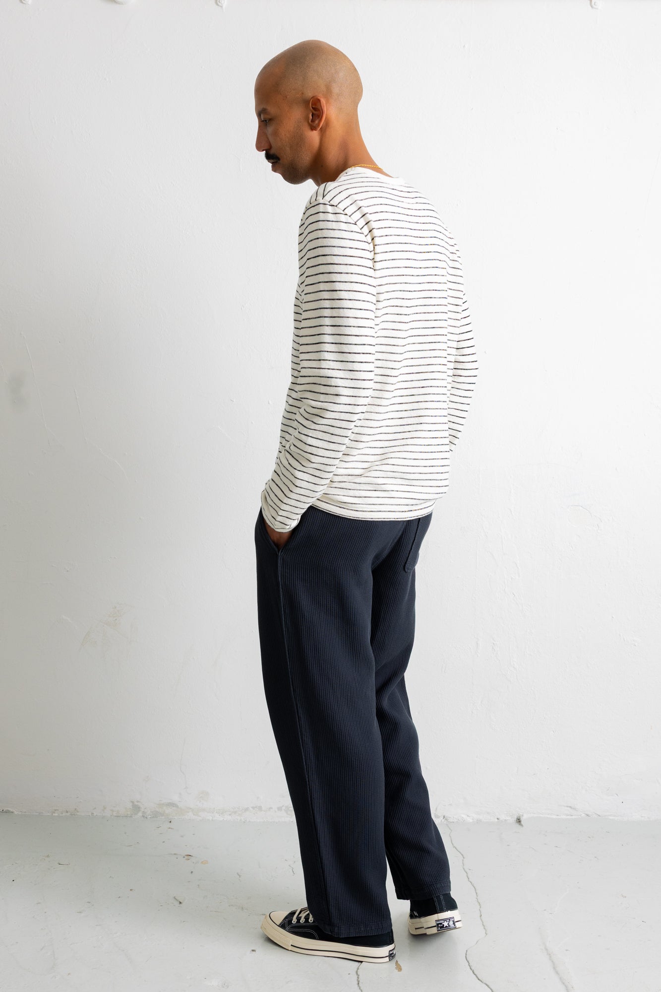 A man wearing a white long-sleeve shirt with thin black horizontal stripes and dark navy wide-leg trousers, standing in profile against a white wall.
