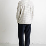 A man viewed from behind wearing a cream-colored long-sleeve shirt with thin dark horizontal stripes, dark navy pants, and black-and-white sneakers, standing against a plain white wall.