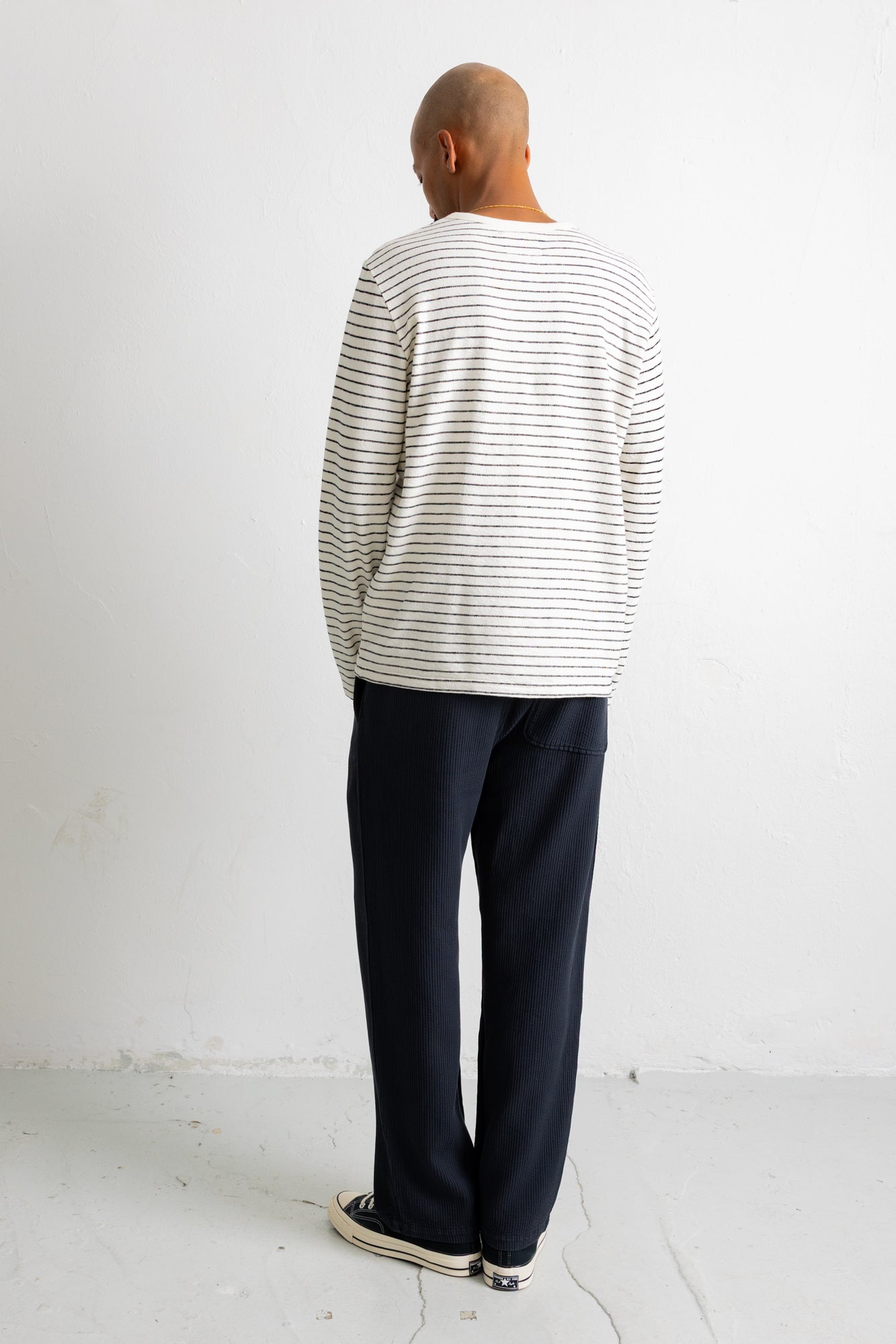 A man viewed from behind wearing a cream-colored long-sleeve shirt with thin dark horizontal stripes, dark navy pants, and black-and-white sneakers, standing against a plain white wall.