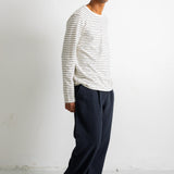 A man wearing a white long-sleeve crewneck shirt with thin dark horizontal stripes, dark navy pants, and black canvas sneakers, standing against a plain white wall.