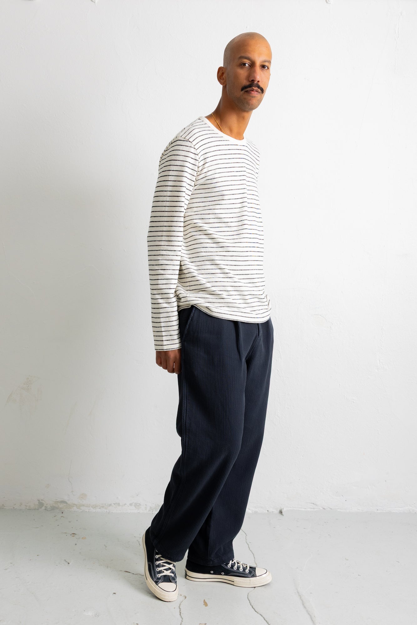 A man wearing a white long-sleeve crewneck shirt with thin dark horizontal stripes, dark navy pants, and black canvas sneakers, standing against a plain white wall.