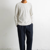 A man wearing a white long-sleeve crewneck shirt with thin dark horizontal stripes, paired with dark navy trousers and black canvas sneakers, standing against a plain white wall.