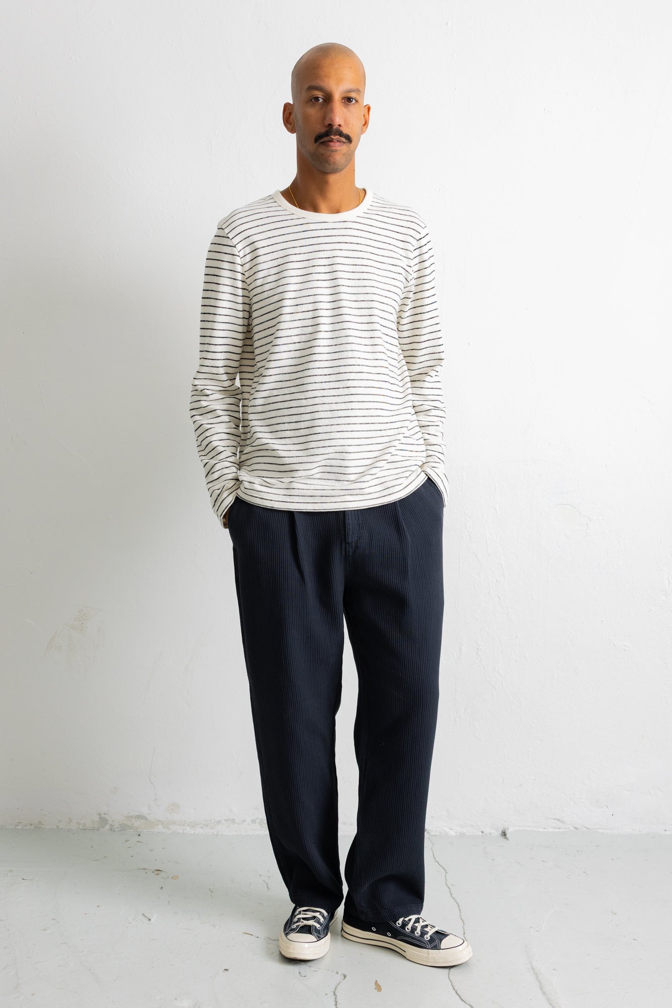 A man wearing a white long-sleeve crewneck shirt with thin dark horizontal stripes, paired with dark navy trousers and black canvas sneakers, standing against a plain white wall.