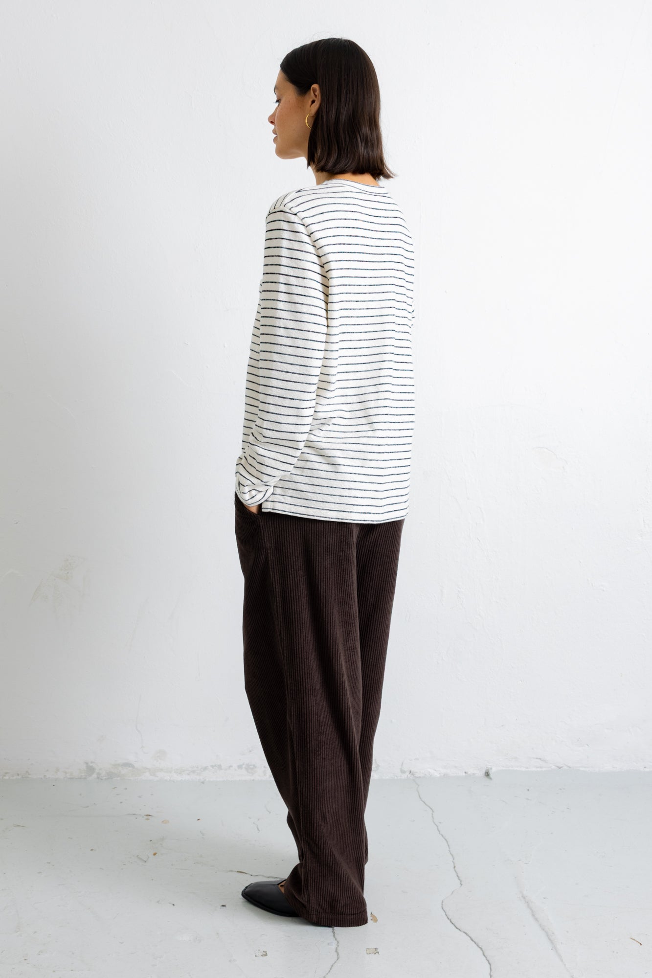 A model shown from the back wearing a white long-sleeve shirt with thin dark horizontal stripes and dark brown wide-leg trousers, standing against a white wall.