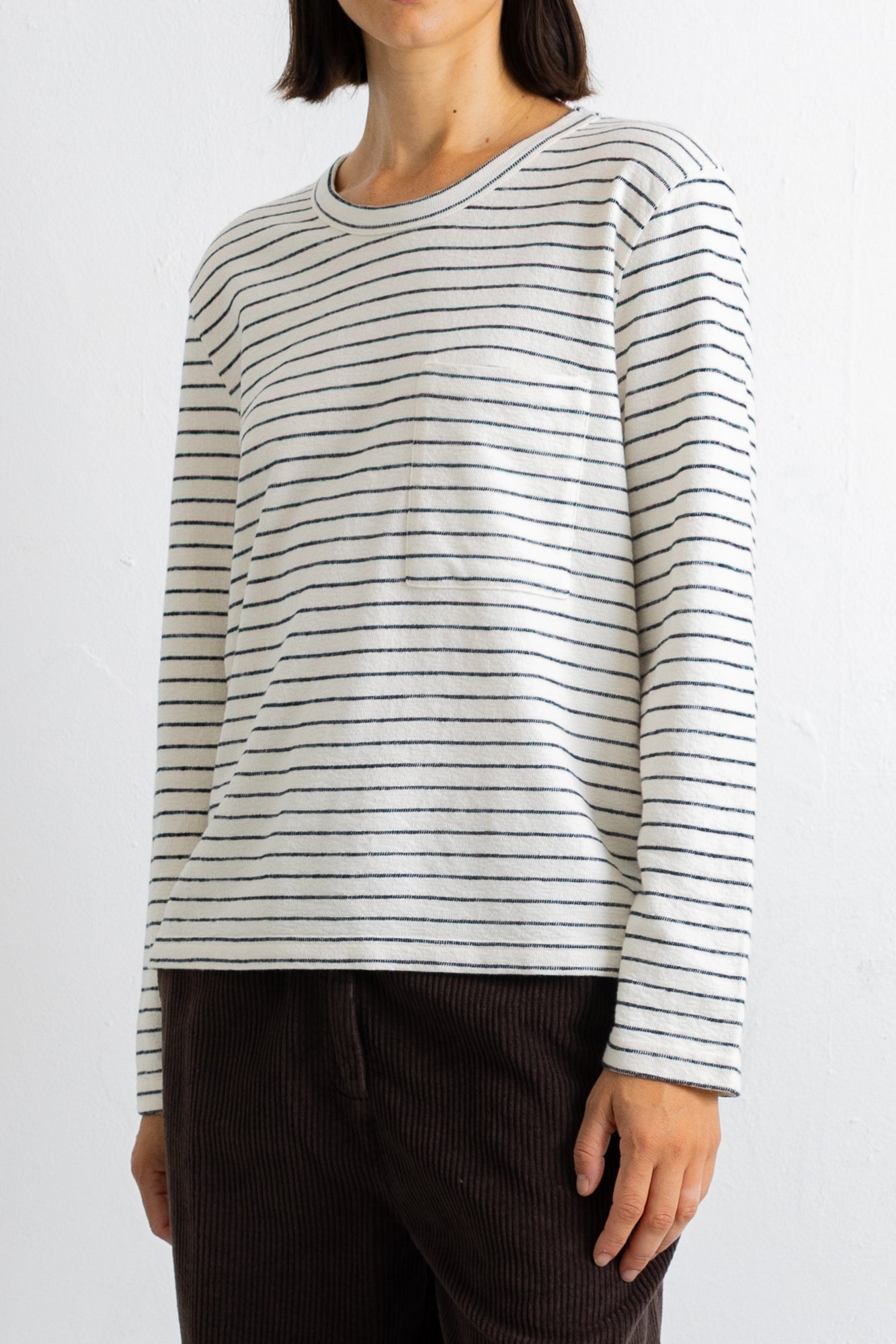 A woman wearing a long-sleeve crewneck shirt with thin dark horizontal stripes on an off-white base, paired with dark brown corduroy pants, against a plain white background.