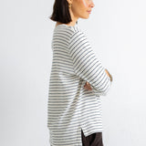 A woman in profile wearing a cream-colored long-sleeve top with thin dark horizontal stripes and dark brown corduroy pants, standing against a plain white wall.