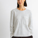 A woman wearing a long-sleeve crewneck top with thin dark horizontal stripes on an off-white base, paired with dark brown corduroy pants, against a plain light gray background.