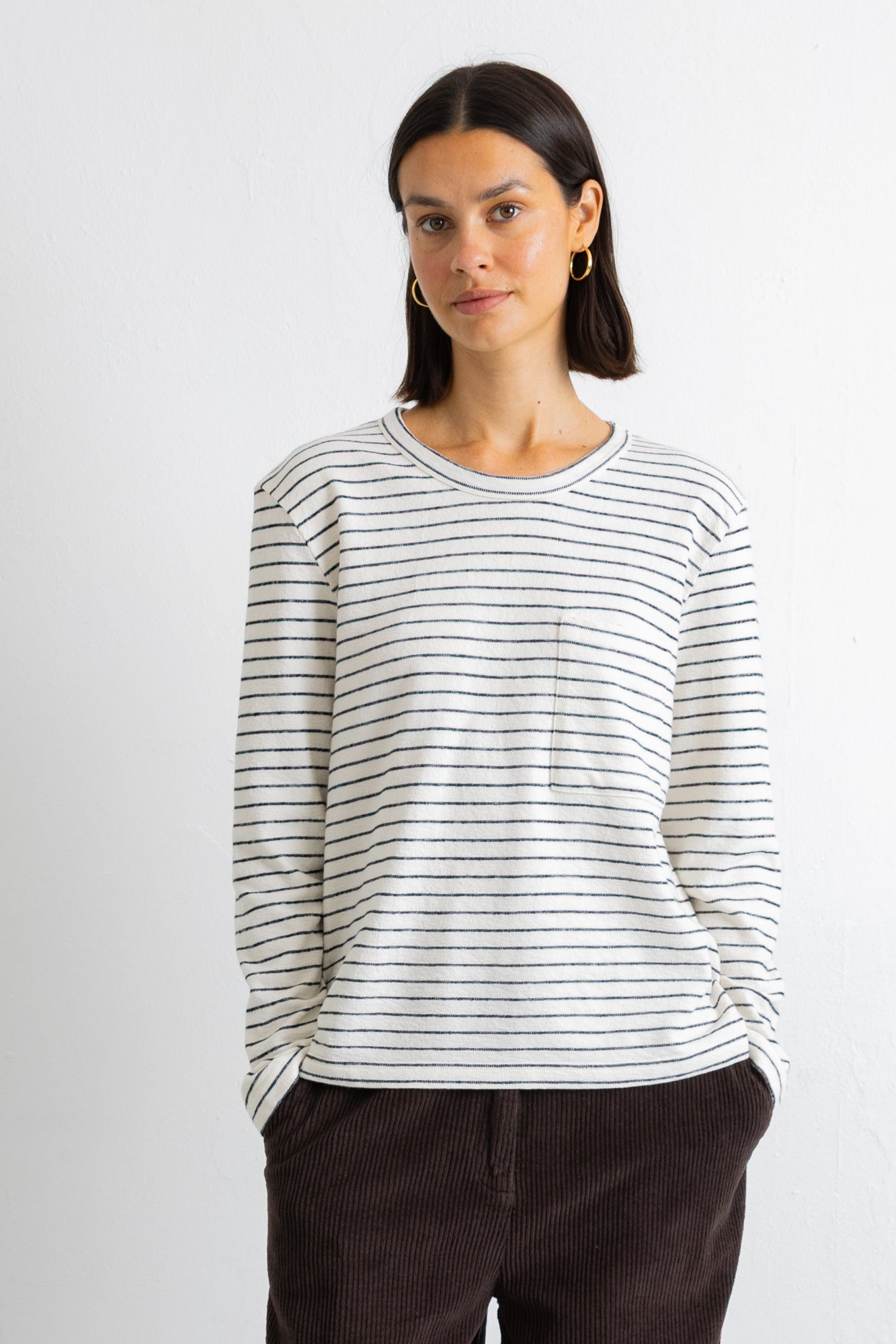A woman wearing a long-sleeve crewneck top with thin dark horizontal stripes on an off-white base, paired with dark brown corduroy pants, against a plain light gray background.