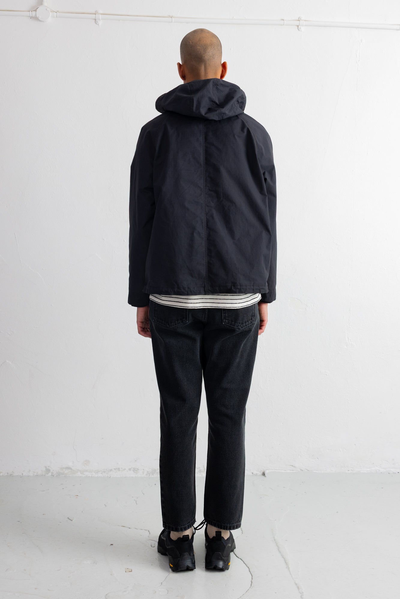 A man viewed from behind wearing a black hooded jacket, black trousers, and black shoes, standing against a plain white wall.