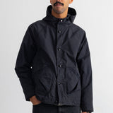 A man wearing a black hooded jacket with snap buttons and large front patch pockets, paired with dark jeans, standing against a plain white studio background.