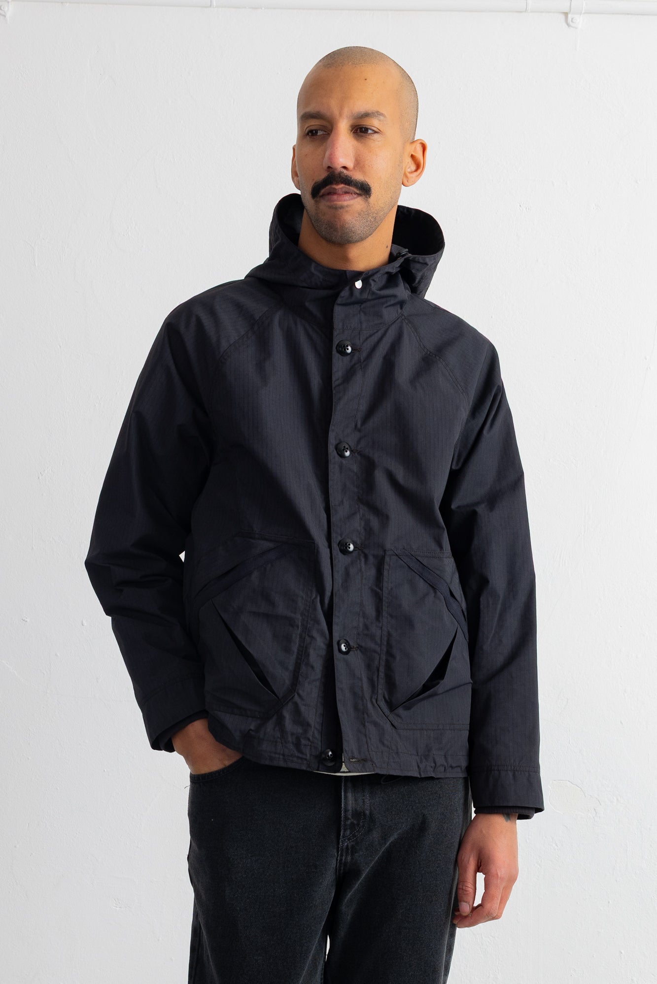 A man wearing a black hooded jacket with snap buttons and large front patch pockets, paired with dark jeans, standing against a plain white studio background.