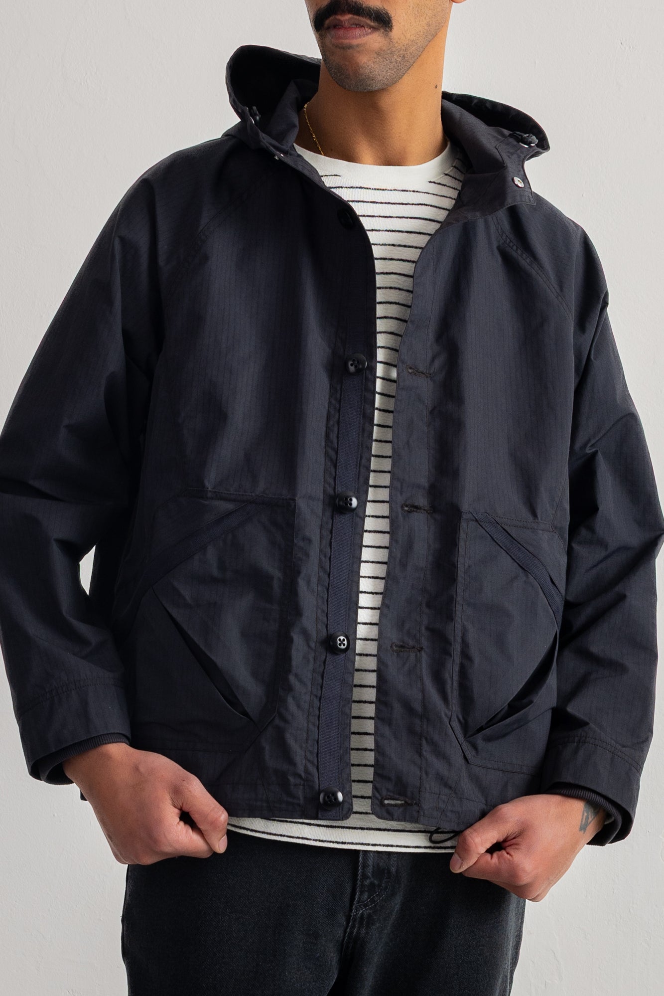 A man wearing a black hooded jacket over a white and gray striped t-shirt, shown from the chin down against a plain light gray background.