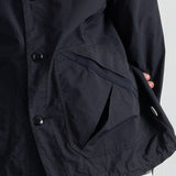 Close-up of a black jacket with a large front flap pocket, black buttons, and a striped shirt peeking out at the hem, shown against a plain light background.