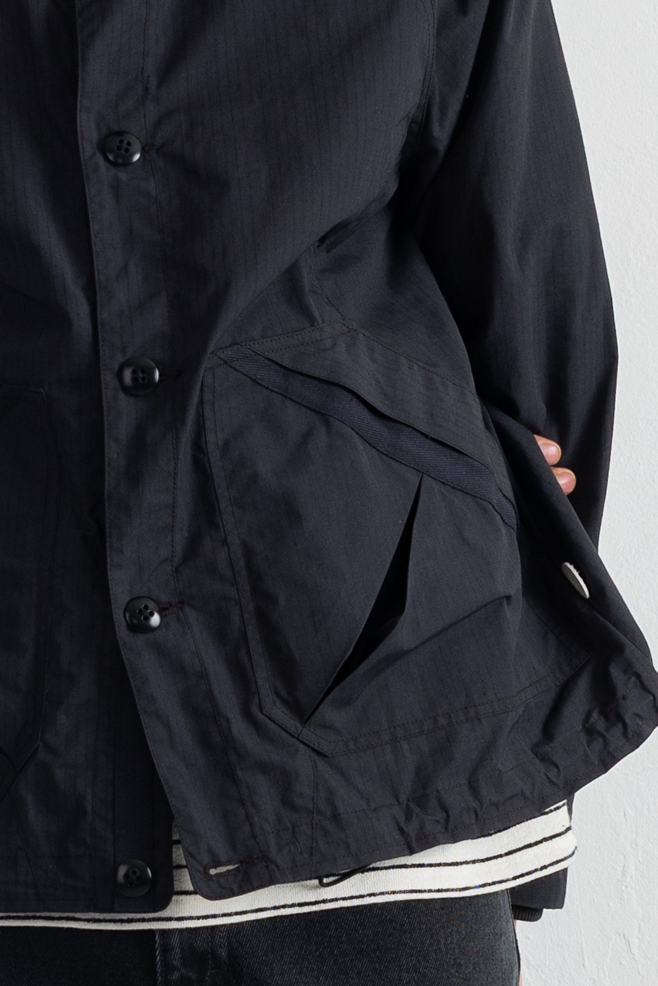 Close-up of a black jacket with a large front flap pocket, black buttons, and a striped shirt peeking out at the hem, shown against a plain light background.