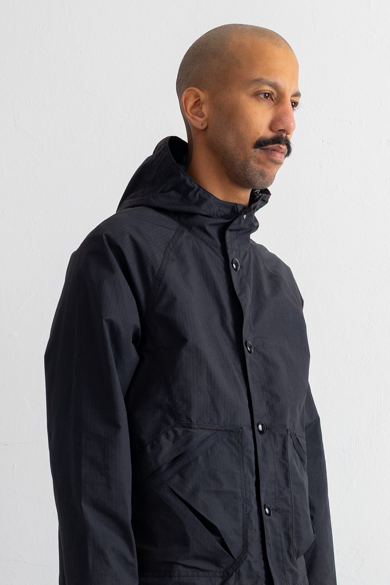 A man wearing a black hooded jacket, shown in a side profile against a plain white wall.