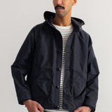 A man wearing a black hooded jacket over a white and gray striped t-shirt, standing against a plain white wall.