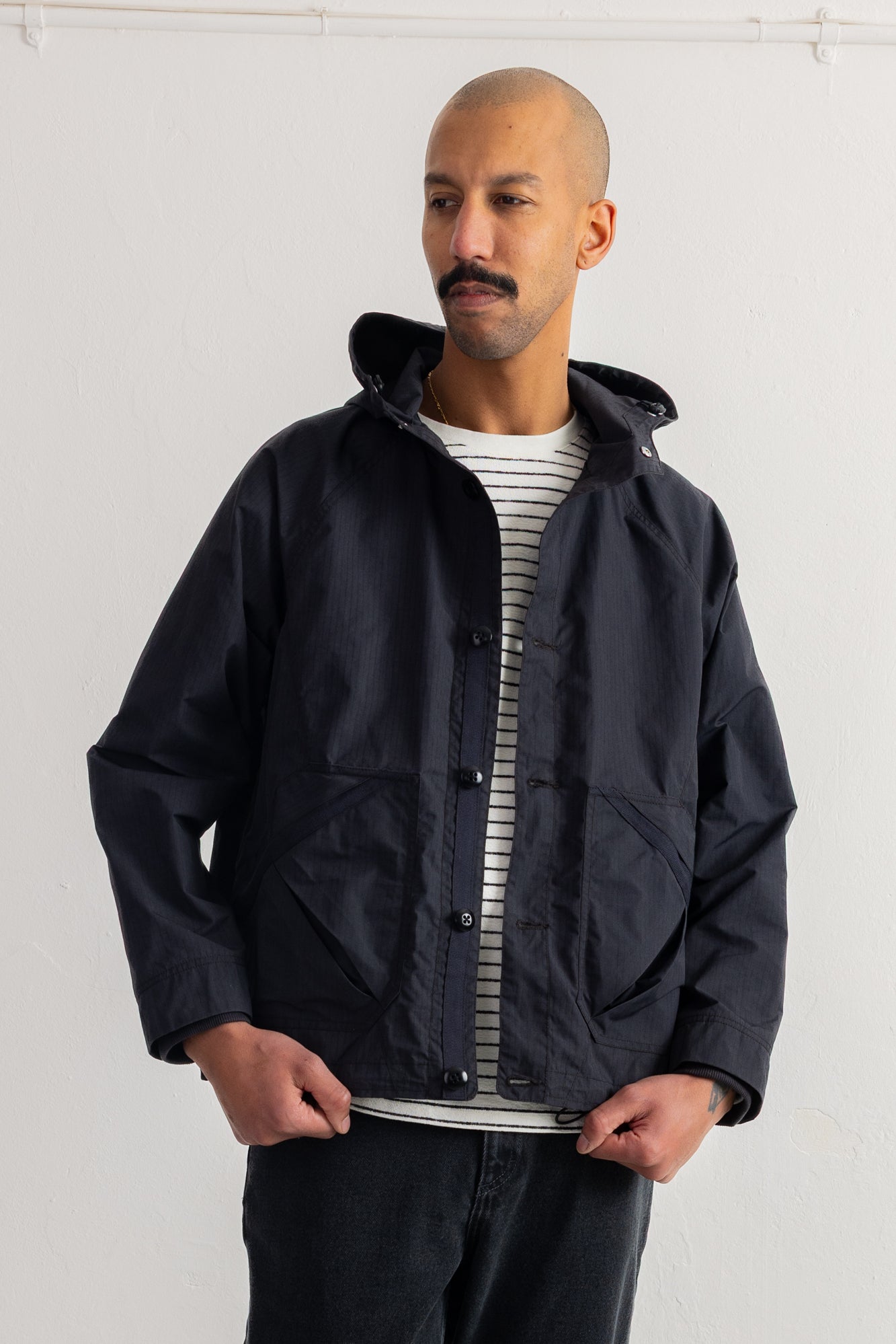 A man wearing a black hooded jacket over a white and gray striped t-shirt, standing against a plain white wall.