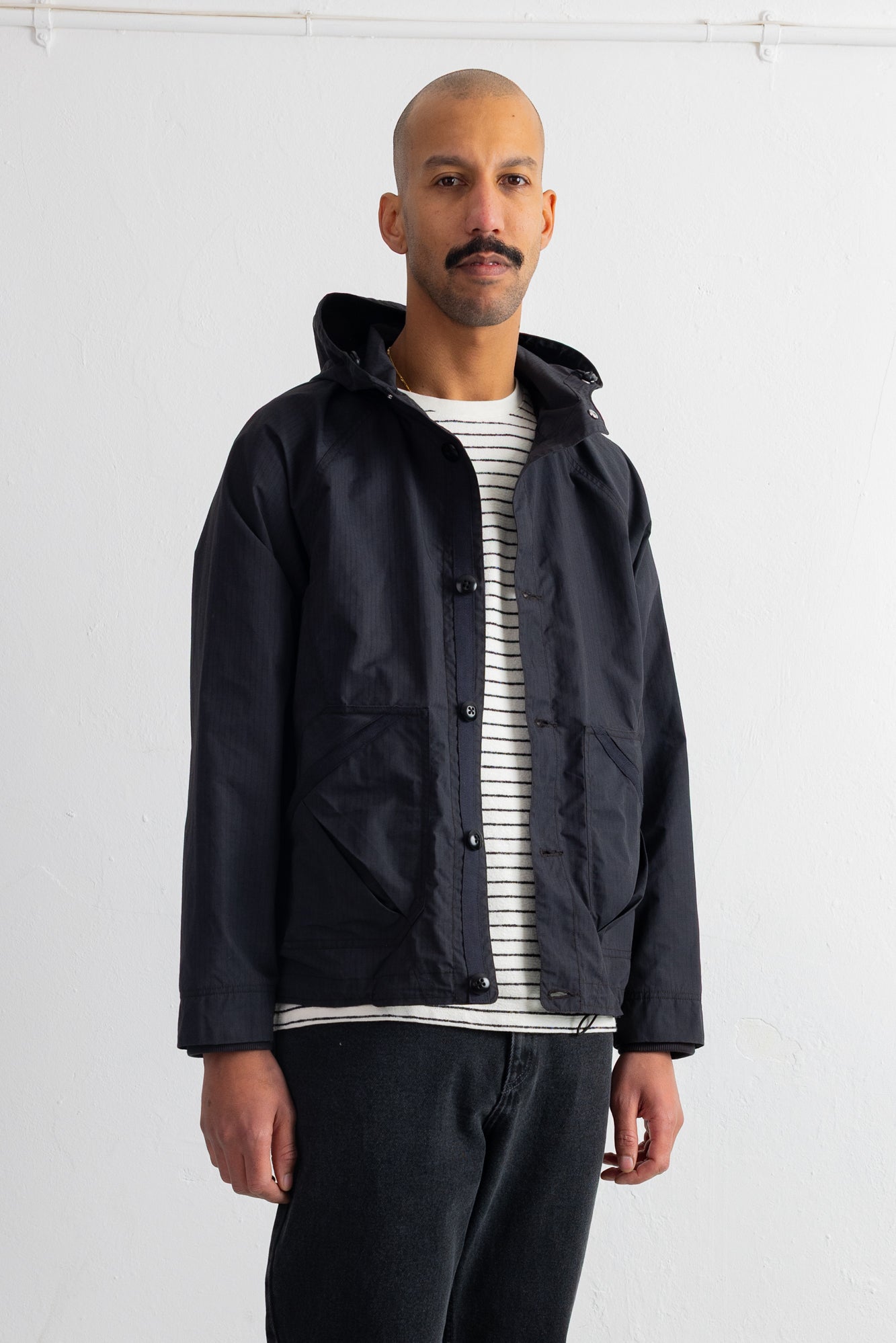 A man wearing a black hooded jacket over a black and white striped shirt, paired with dark jeans, standing against a plain white wall.