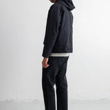 A man in profile wearing a black hooded jacket, black jeans, and black sneakers, standing against a plain white studio wall.