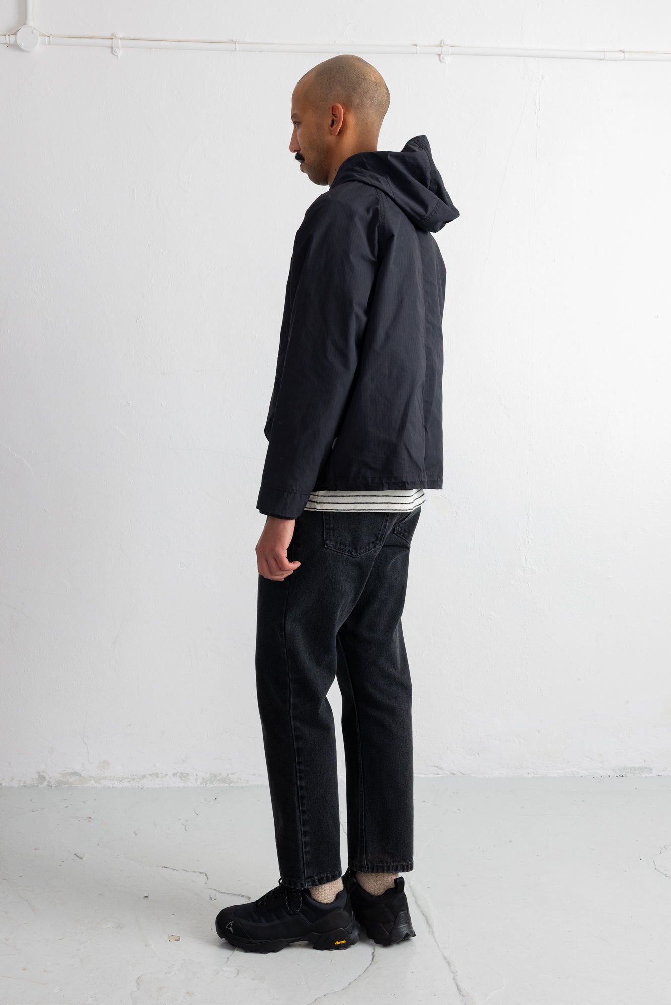 A man in profile wearing a black hooded jacket, black jeans, and black sneakers, standing against a plain white studio wall.