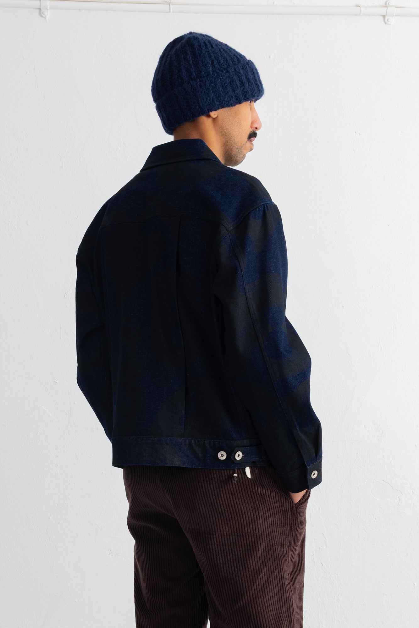 A man wearing a dark navy blue jacket, a matching navy blue knit beanie, and brown corduroy trousers, photographed from the back against a plain white wall.