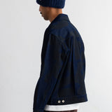 A person shown in side profile wearing a dark navy textured knit beanie, a dark navy jacket over a white shirt, and brown corduroy trousers, against a plain white studio background.