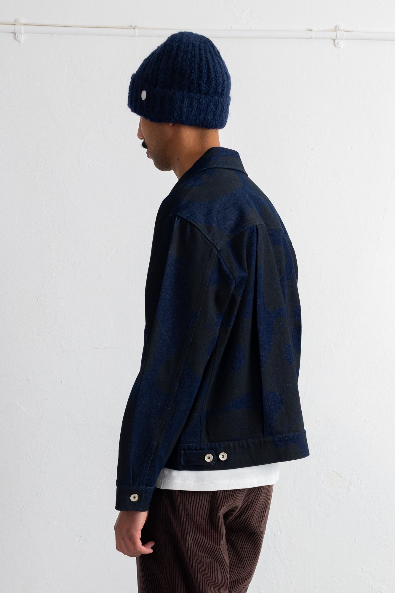 A person shown in side profile wearing a dark navy textured knit beanie, a dark navy jacket over a white shirt, and brown corduroy trousers, against a plain white studio background.