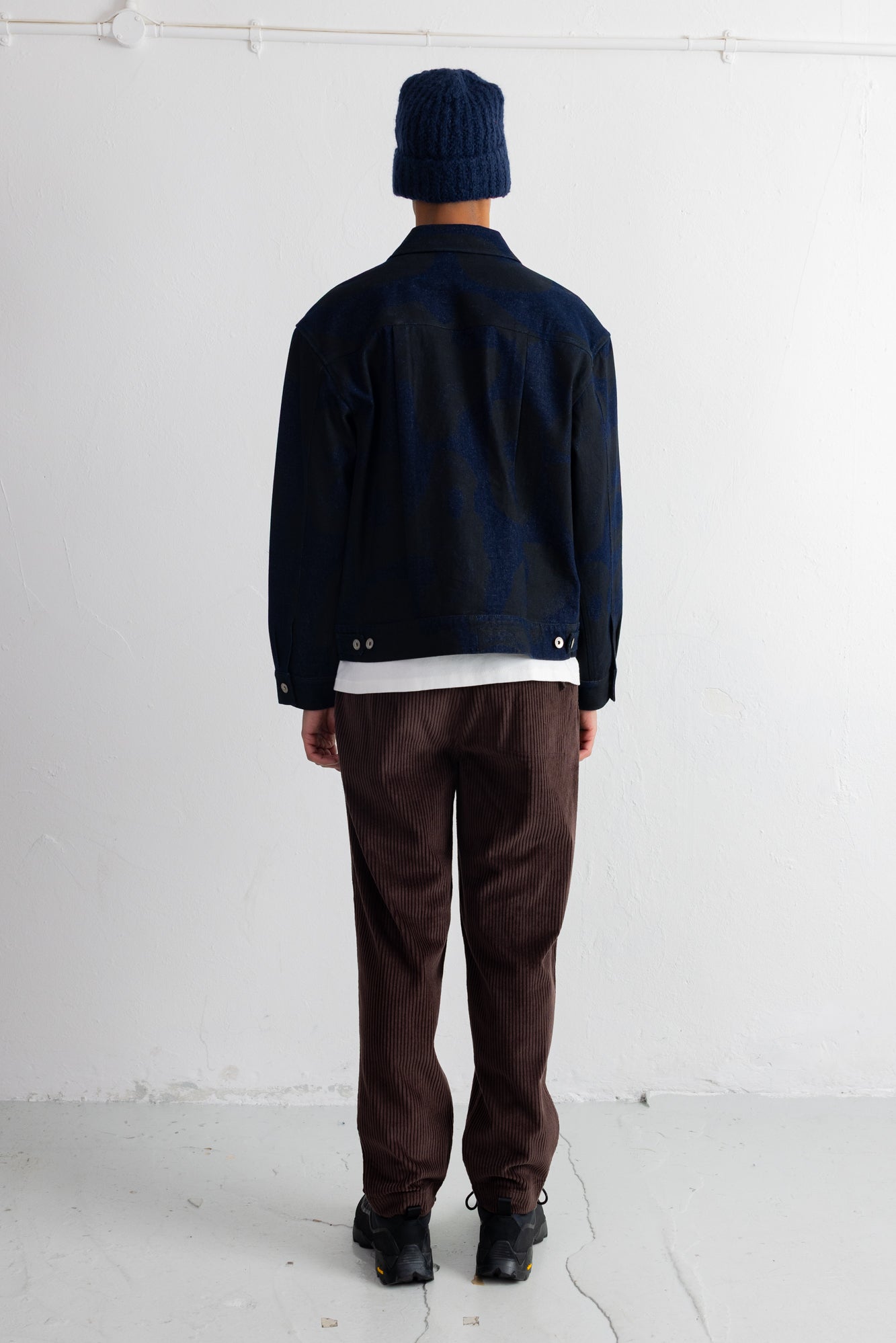 A person shown from the back wearing a dark navy jacket, dark brown pants, a dark navy beanie, and black shoes, standing against a plain white wall.