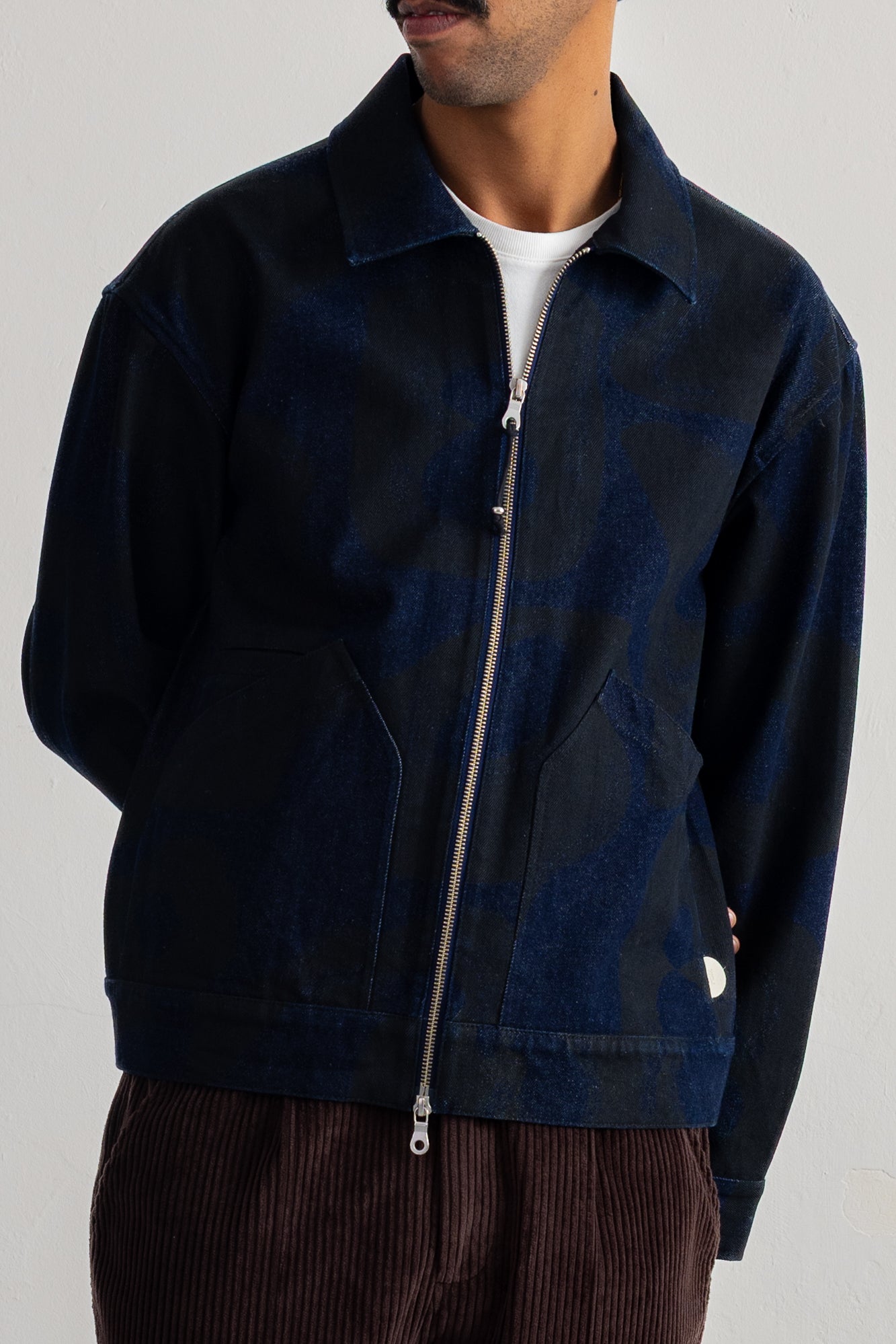 A man wearing a dark navy blue zip-up jacket with a subtle tonal pattern, layered over a white t-shirt and brown corduroy trousers, photographed against a neutral light gray background.