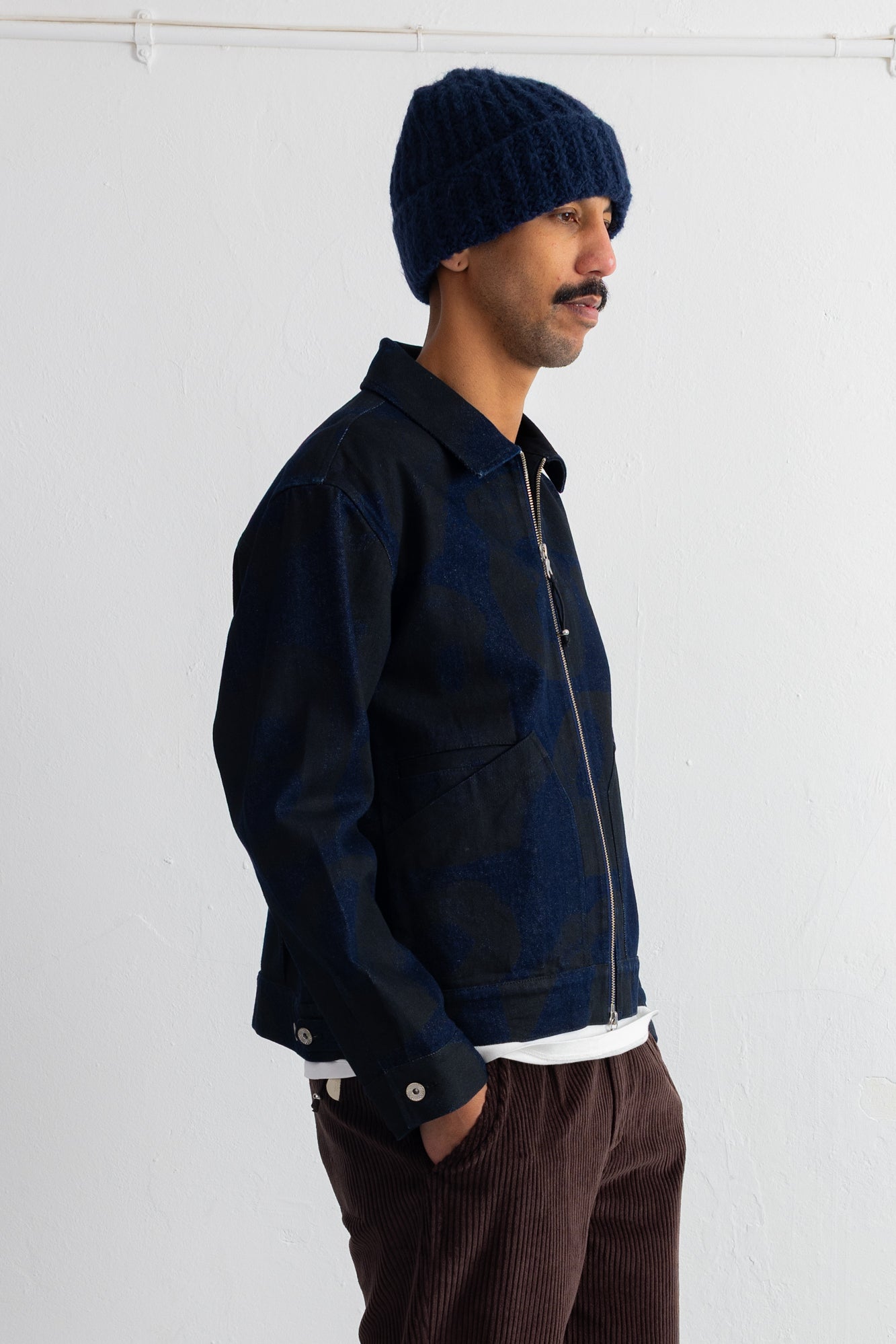 A man wearing a dark navy blue zip-up jacket, a matching navy knit beanie, and brown corduroy pants, shown in profile against a plain white wall.