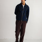 A man wearing a dark navy blue zip-up jacket, dark brown corduroy pants, a navy blue knit beanie, and black shoes, standing against a plain white studio wall.