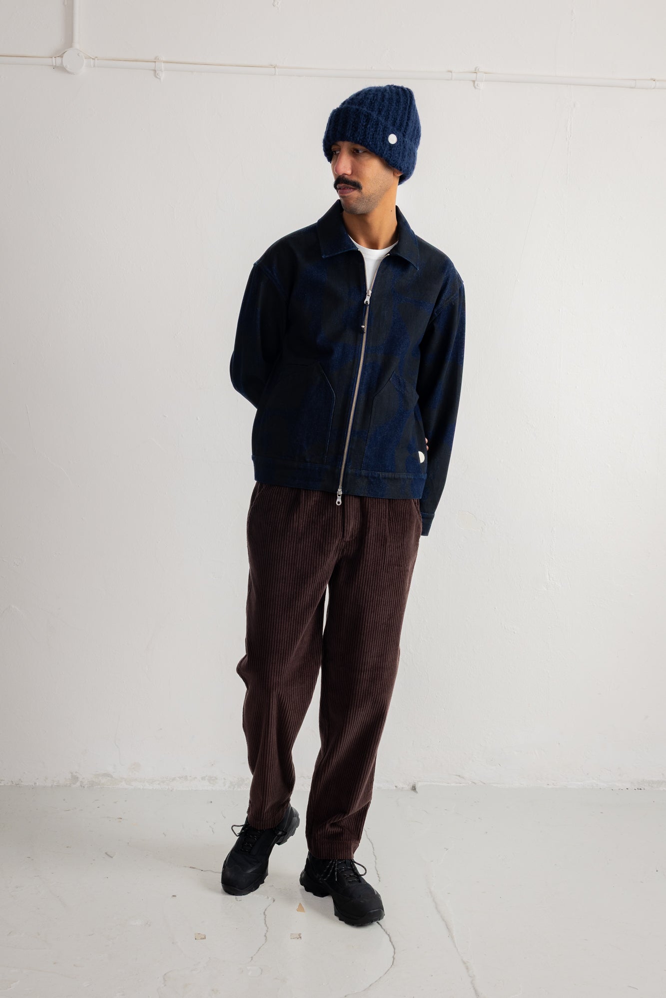 A man wearing a dark navy blue zip-up jacket, dark brown corduroy pants, a navy blue knit beanie, and black shoes, standing against a plain white studio wall.