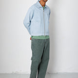 Man wearing a light blue jacket and green pants standing against a white wall.