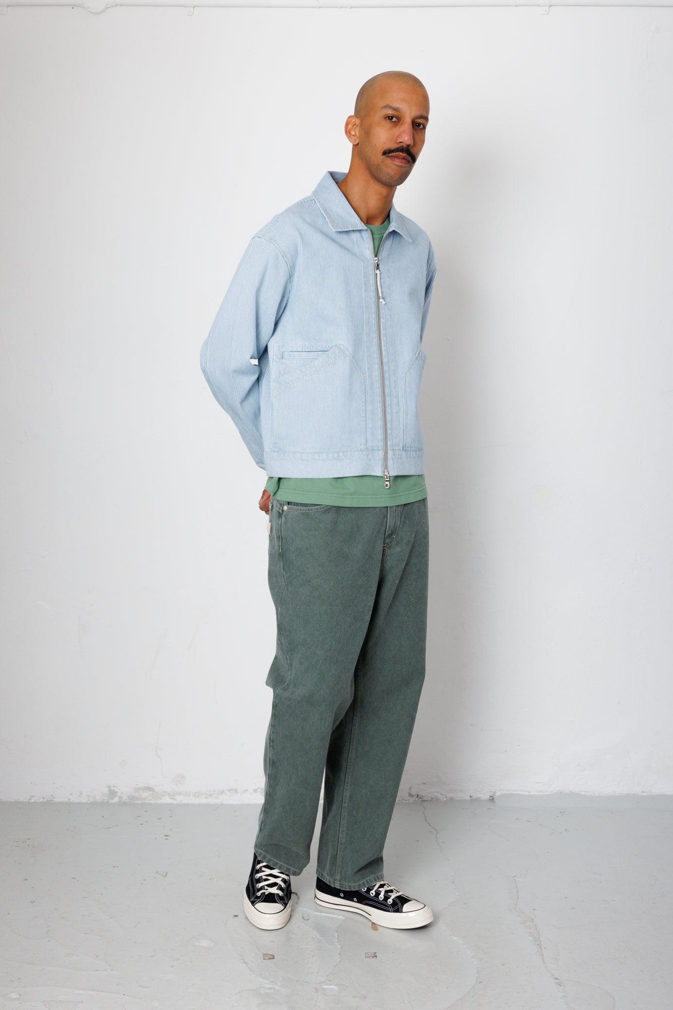 Man wearing a light blue jacket and green pants standing against a white wall.